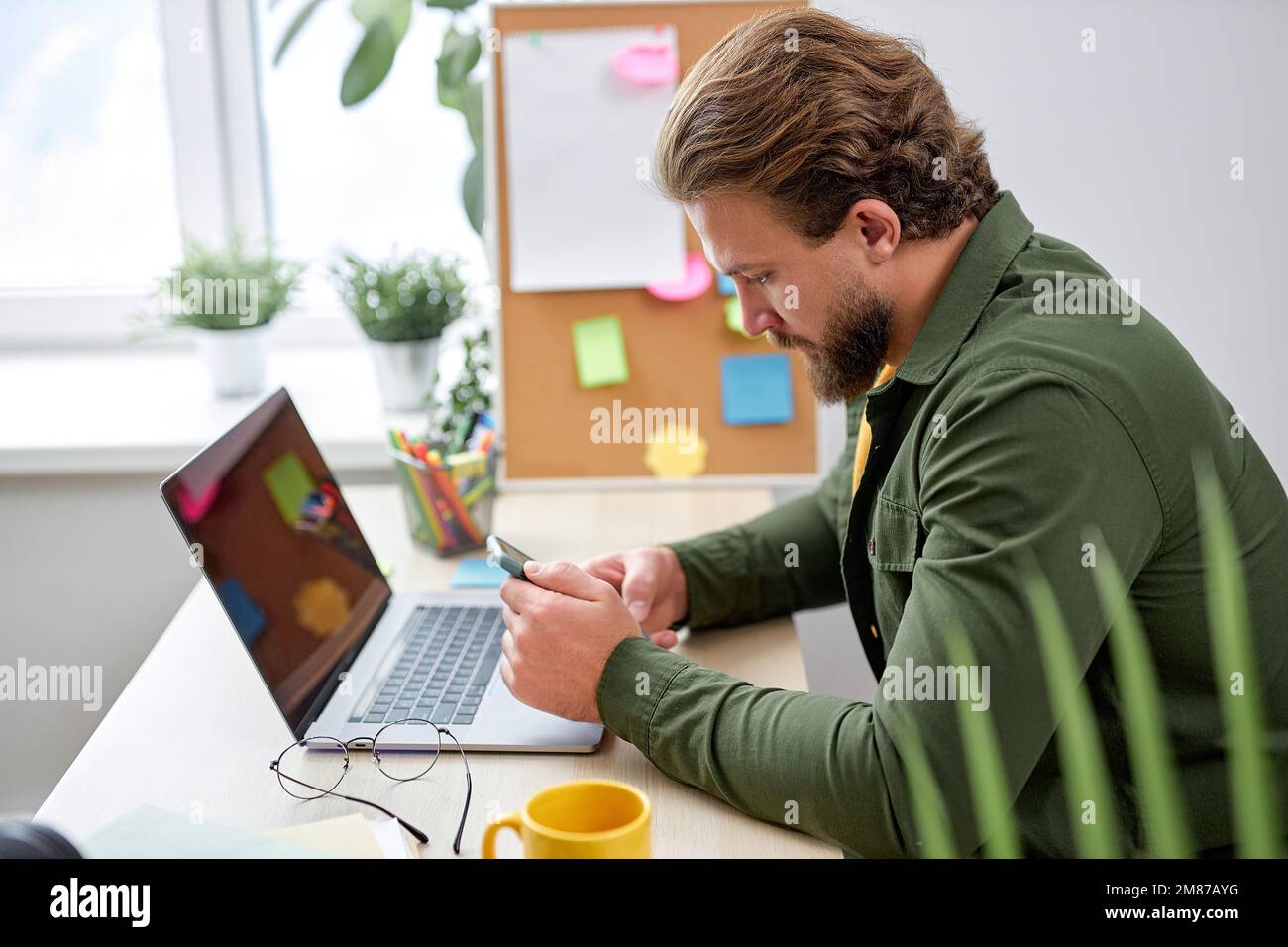 Der junge Mann sitzt mit dem Smartphone und ruht sich während der Arbeit in einem hellen, modernen Büro aus, allein. Ein bärtiger weißer Kerl in Freizeitkleidung macht eine Pause beim Tippen von Messa Stockfoto