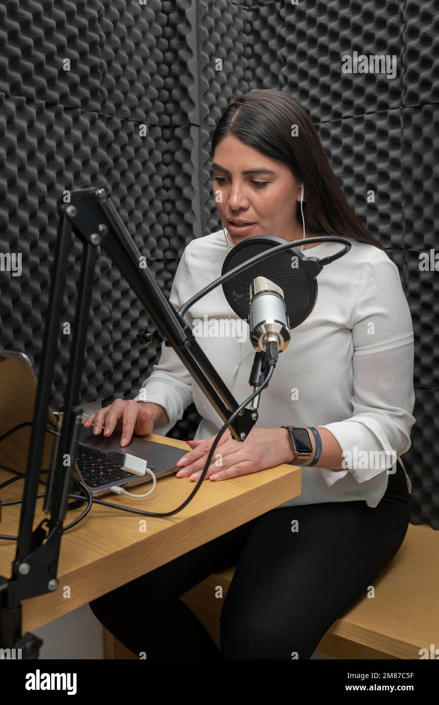 Frau spricht in ein Mikrofon, während sie einen Podcast aufnimmt, und verwendet dabei eine Audiokabine mit akustischer Isolierung. Stockfoto