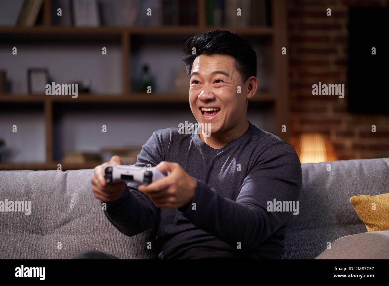 Glücklicher asiatischer Mann, der einen drahtlosen Joystick verwendet und zu Hause Videospiele spielt. Fröhlicher Mann, der PlayStation nutzt und in virtuellen Spielen gegeneinander antreten kann, während er sich ausruht Stockfoto