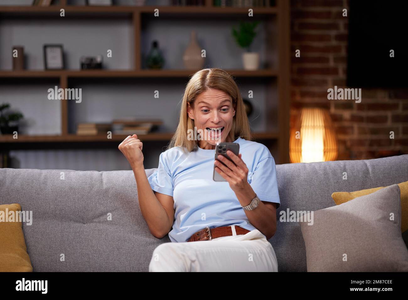 Frau feiert den Sieg, indem sie auf dem Sofa mit Smartphone Ja-Geste zeigt. Wir waren überglücklich, zu gewinnen, lasen die guten Nachrichten am Telefon und feierten den Erfolg unter Stockfoto