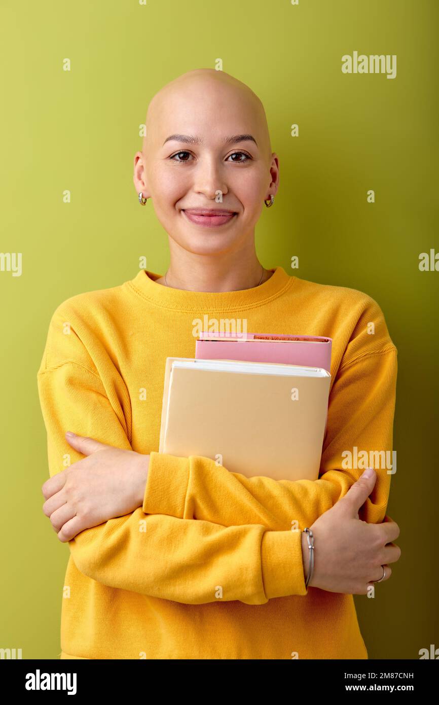 Kahlköpfige, junge Kaukasierin, die auf die Kamera schaut und Bücher in der Hand hält, Alopezie und Krebskonzept, Kopierraum. Zufriedene, haarlose Frau in Casu Stockfoto