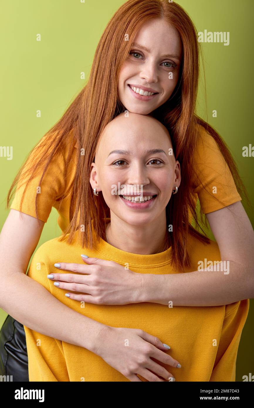 Zwei aufgeregte Damen mit rotem und kahlköpfigem Kopf, die in die Kamera schauen, sich umarmen, isoliert vor grünem Studiohintergrund. Porträt von fröhlich Stockfoto