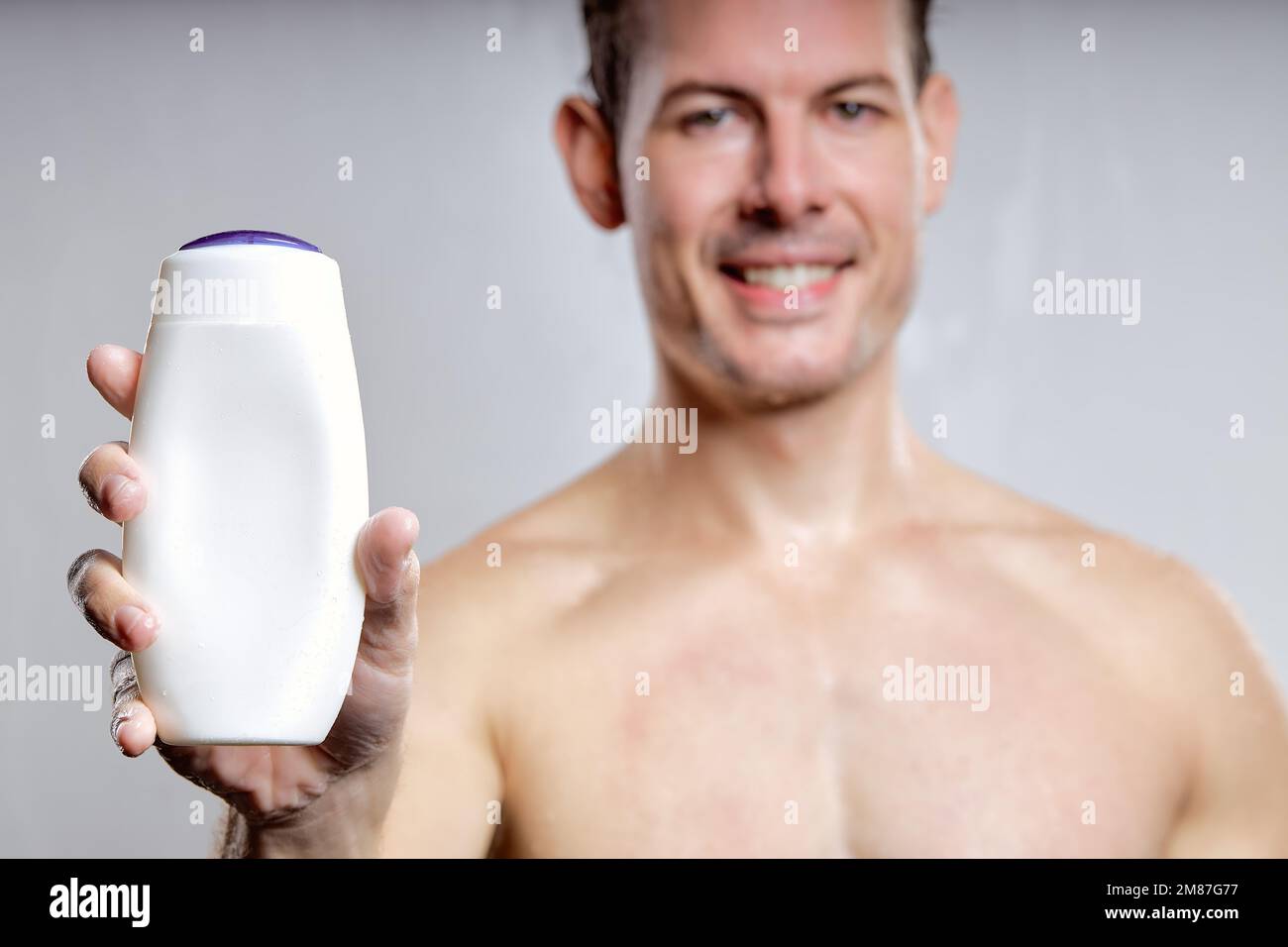 Nahaufnahme eines aufgeregten Mannes, der eine Flasche Gel oder Shampoo zeigt, zur Werbung, isoliert auf grauem Studiohintergrund. Junger Mann mit weißem Notenblatt Stockfoto