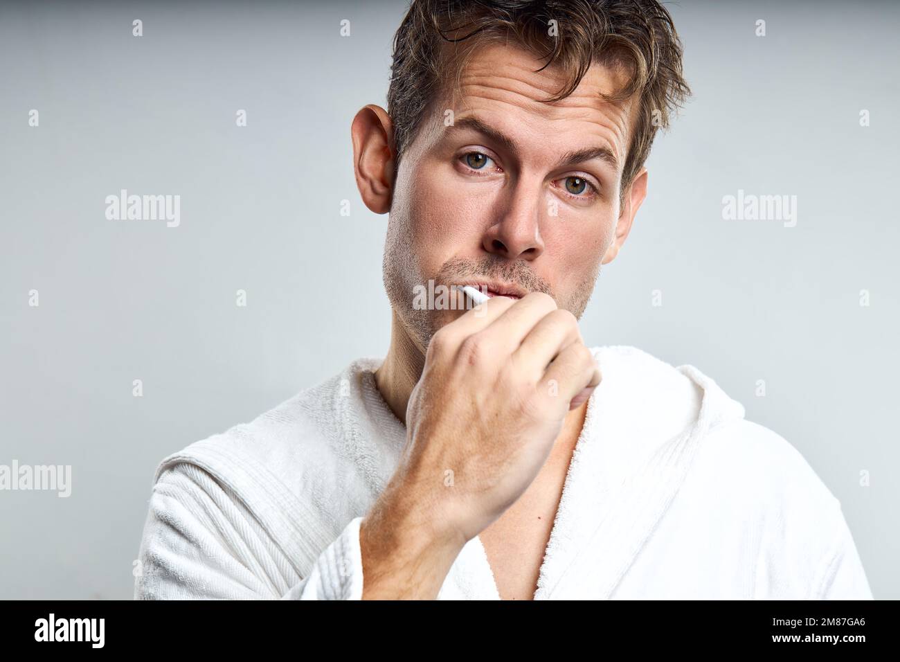 Verwirrt Kerl Zähneputzen in den frühen Morgen, nach dem Aufwachen. Isoliert auf grau. kaukasischen Mann in weißem Bademantel Blick auf die Kamera mit frustrierten Gesichts Stockfoto