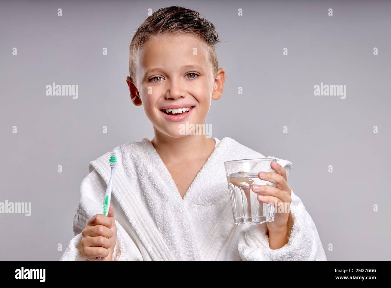 Lächelnde kinderhaltende Zahnbürste und Glas Wasser in den Händen, Zahnhygiene und Gesundheit für Kinder. Plastikkind im weißen Bademantel Genießen Sie das morgendliche Rou Stockfoto