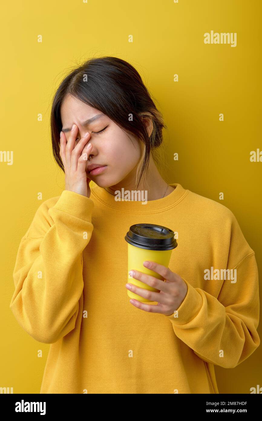 Frustrierte asiatische Frau leidet unter Kopfschmerzen, berührt das Gesicht, trinkt Kaffee nach schlafloser Nacht, hält Einwegbecher, trägt stilvolles lässiges Yello Stockfoto