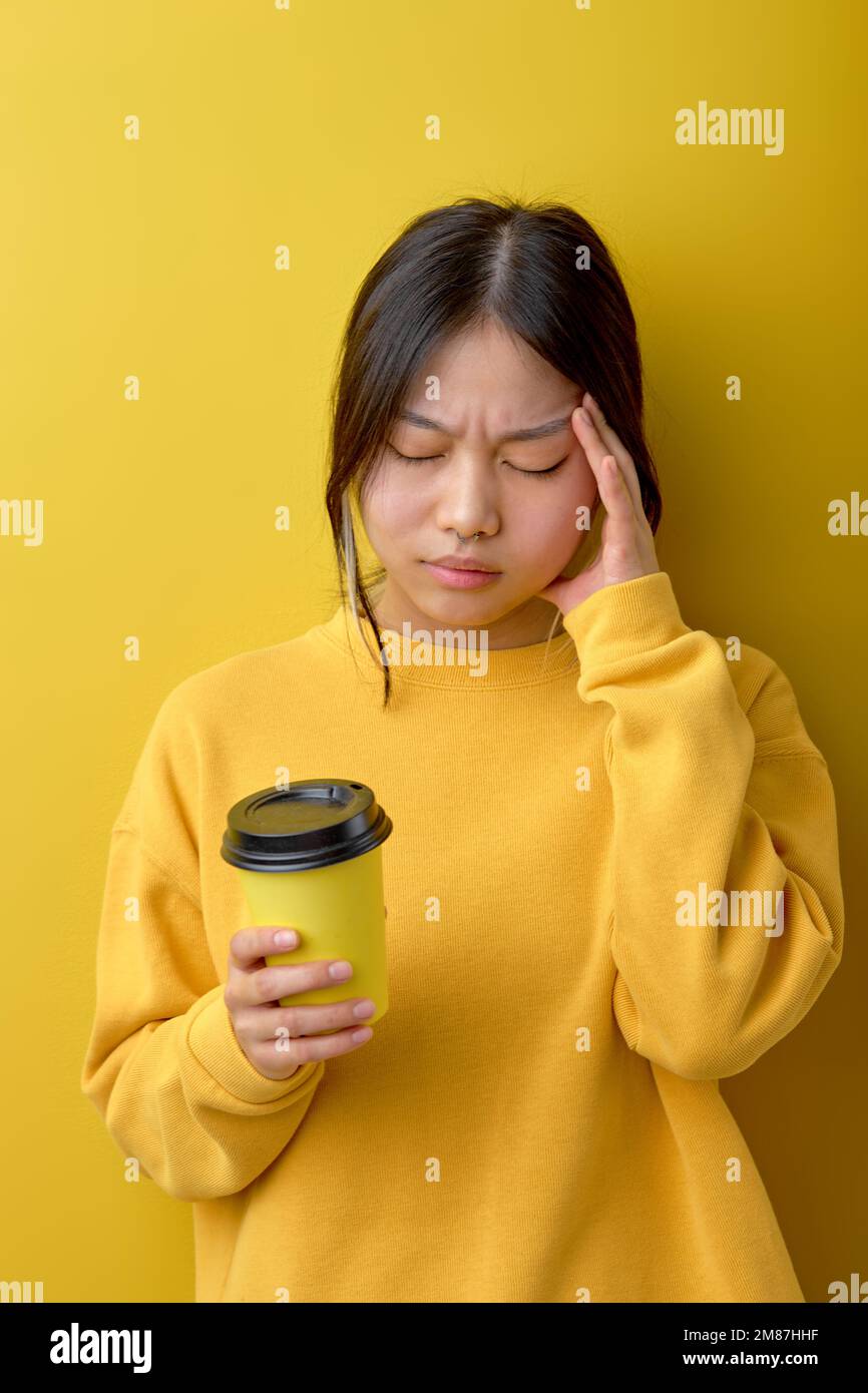 Frustrierte asiatische Frau leidet unter Kopfschmerzen, berührt Tempel, trinkt erfrischendes Getränk nach schlafloser Nacht, hält Einweg-Tasse Kaffee, dresse Stockfoto