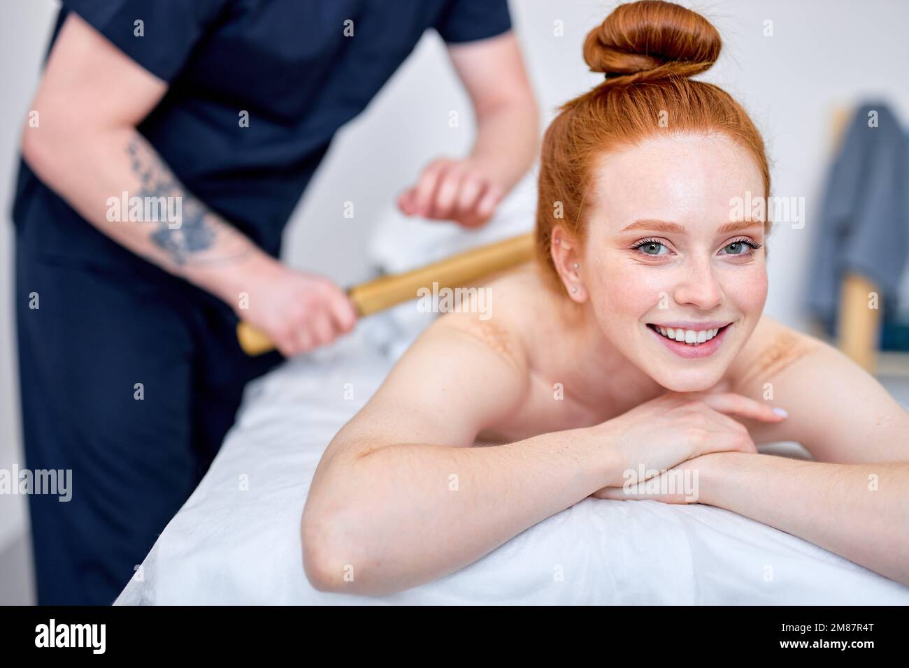 Unerkennbarer professioneller Masseur massiert den Rücken eines zufriedenen Kunden mit Bambusstäbchen, Fokus auf rothaarige, weibliche Klientin, die auf der Couch liegt, lächelt, ha Stockfoto