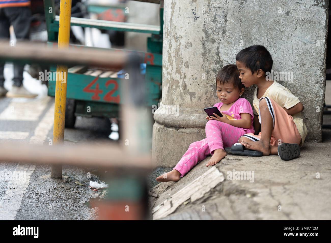 Ein kleines Kind auf den Philippinen sitzt auf einer Seitenstraße mit einem Handy, während ein anderer zusieht. Stockfoto