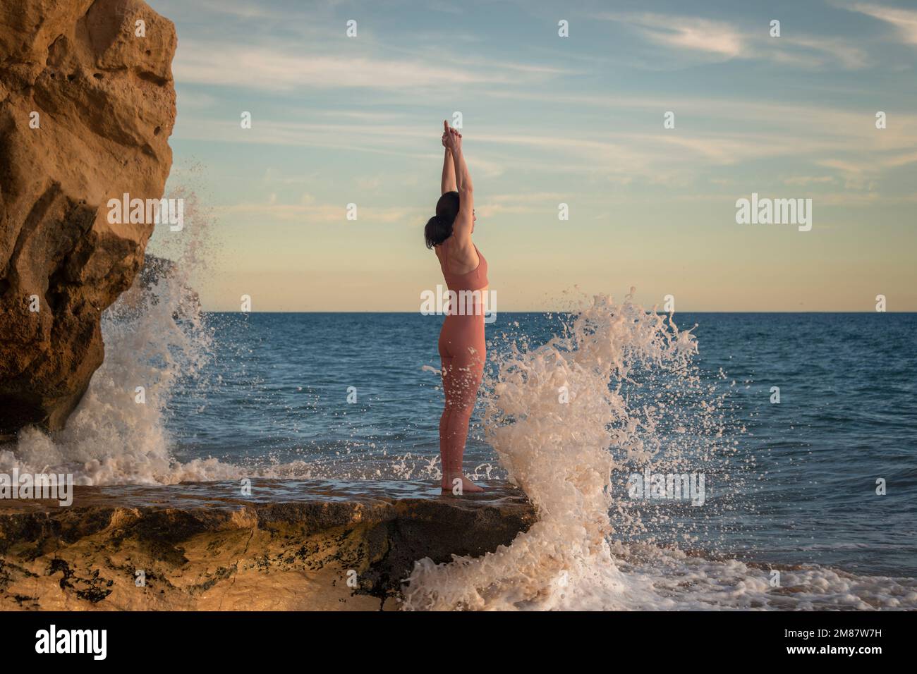 Eine Frau, die auf Felsen am Meer steht, Wellen brechen, Arme hoch beim Yoga üben. Stockfoto