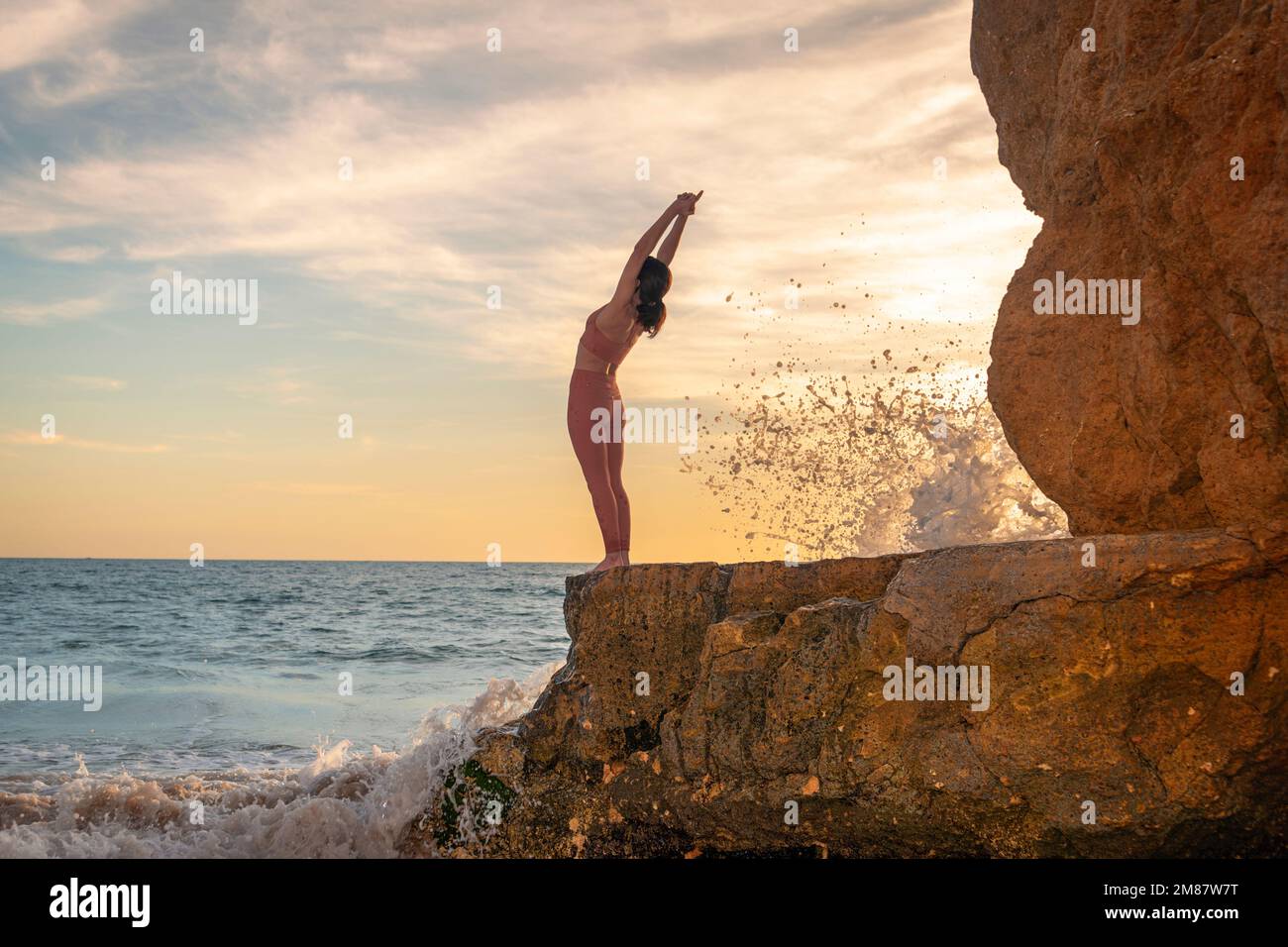 Eine Frau, die auf Felsen am Meer steht und eine Yoga-Pose macht. Stockfoto