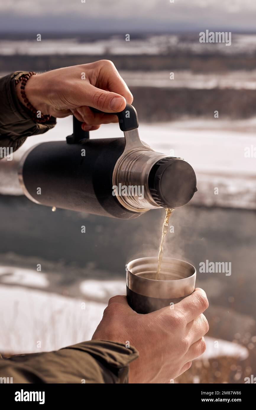 Ein unbekannter männlicher Reisender, der in der verschneiten Winterlandschaft aus der Thermoskanne heißen Tee in die Tasse gießt. bushcraft-Konzept. Nahaufnahmen, gekürzter Mann, Fluss in der Stockfoto