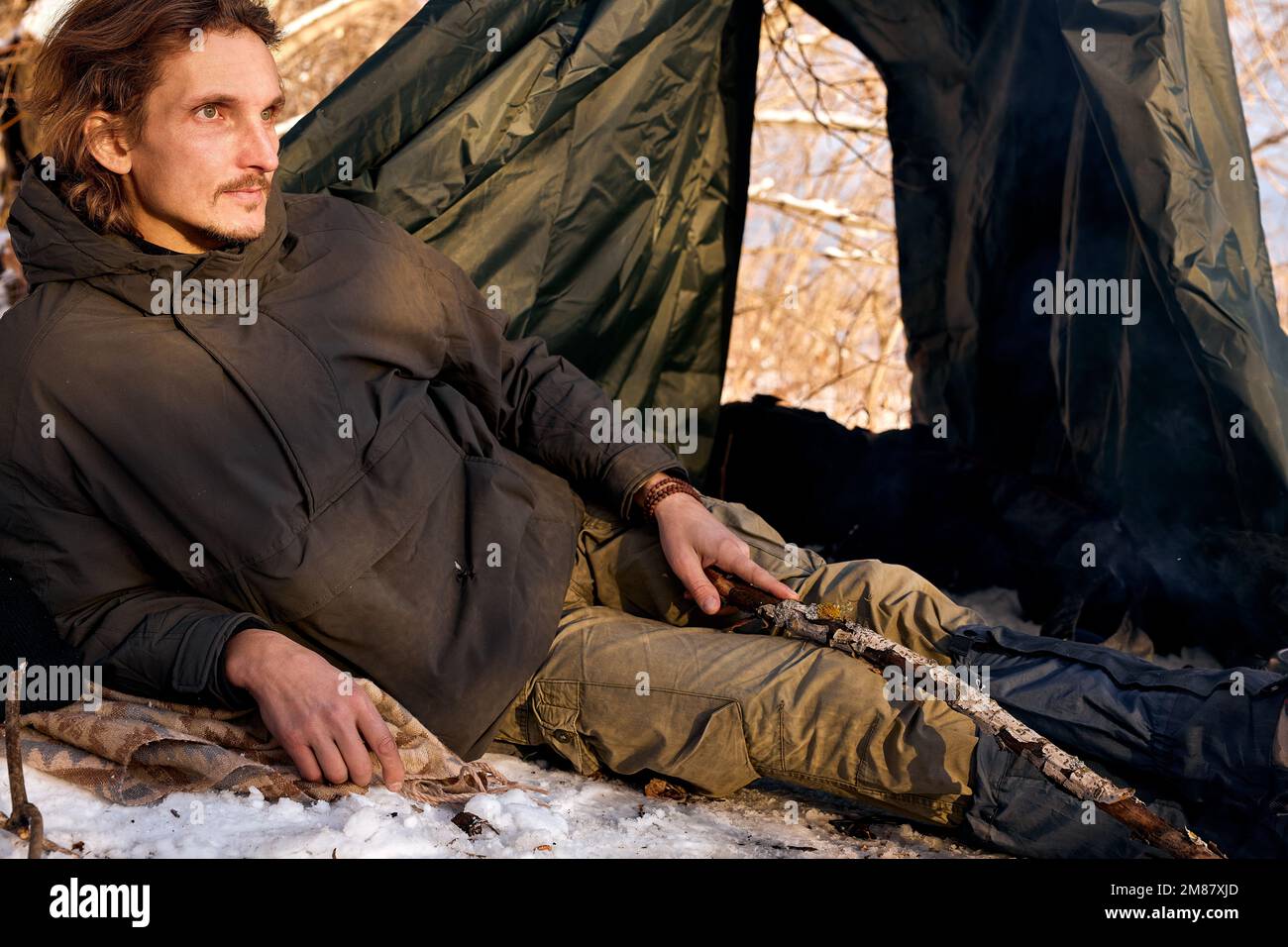 Ruhiger entspannter kaukasischer Kerl in warmer Kleidung liegend neben dem Zelt während des Campens, allein. Junge Männchen haben Ruhe, entspannend, Blick auf Seite in Kontemplation, en Stockfoto