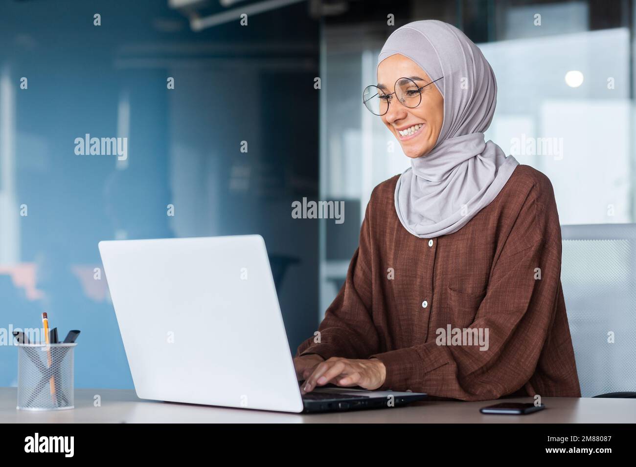 Junge, wunderschöne muslimische Frau im Hijab, die in einem modernen Büro arbeitet, Geschäftsfrau, die ein Notebook bei der Arbeit verwendet, Tastatur eingibt, während sie am Arbeitsplatz sitzt und lächelt und mit dem Erfolg zufrieden ist. Stockfoto