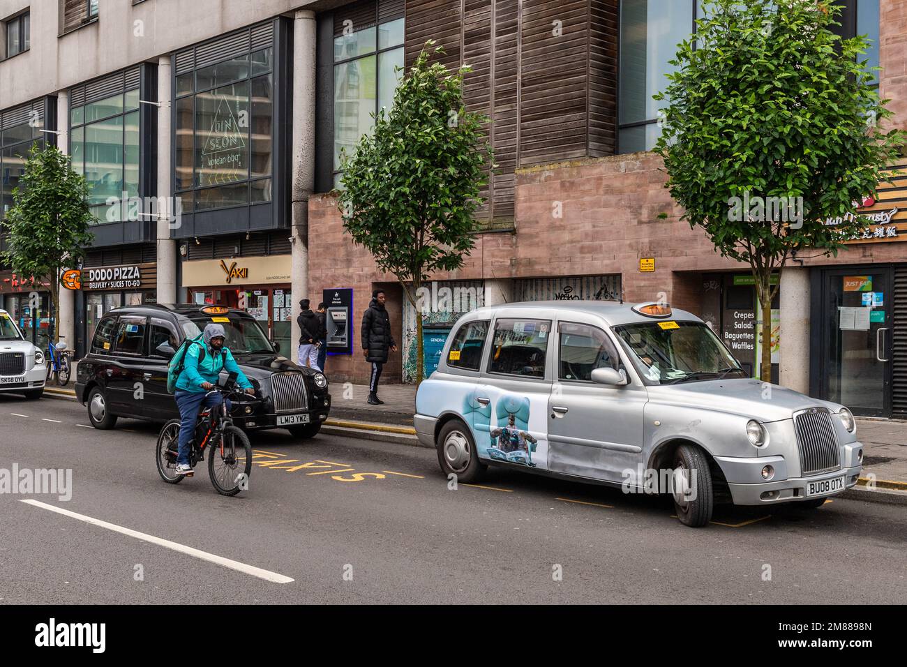 Männlicher Radfahrer von Deliveroo, der in Coventry, West Midlands, Großbritannien, an den gemieteten Taxis vorbeifährt. Stockfoto
