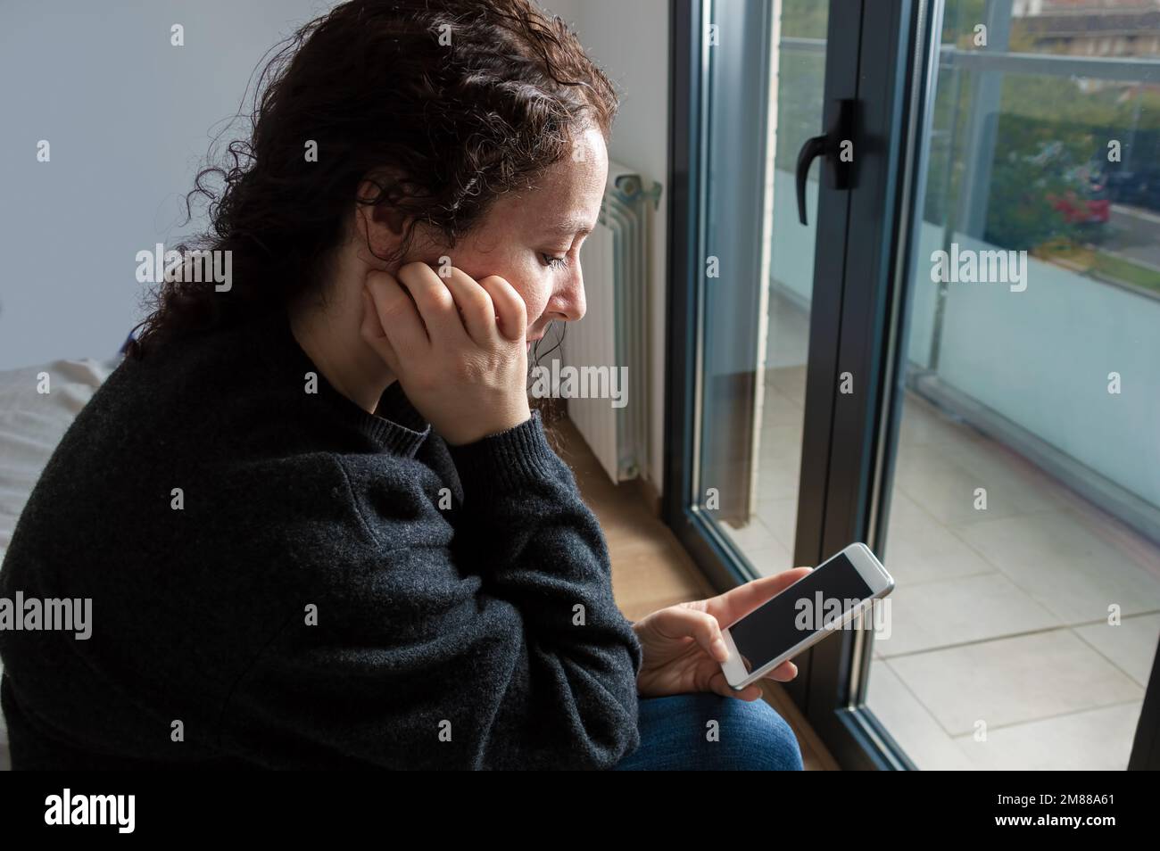 Seitenansicht einer traurigen Frau, die das Telefon überprüft und auf dem Bett im Schlafzimmer neben einem Fenster sitzt Stockfoto