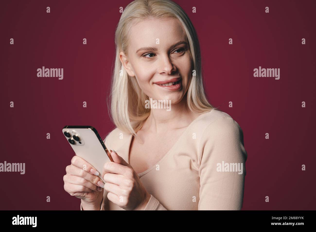Porträt einer hübschen Blondine, die auf einem kirschfarbenen Hintergrund steht und am Telefon SMS schreibt, mit einem nachdenklichen Gesichtsausdruck, der ihr auf die Lippen beißt. Konzept Stockfoto