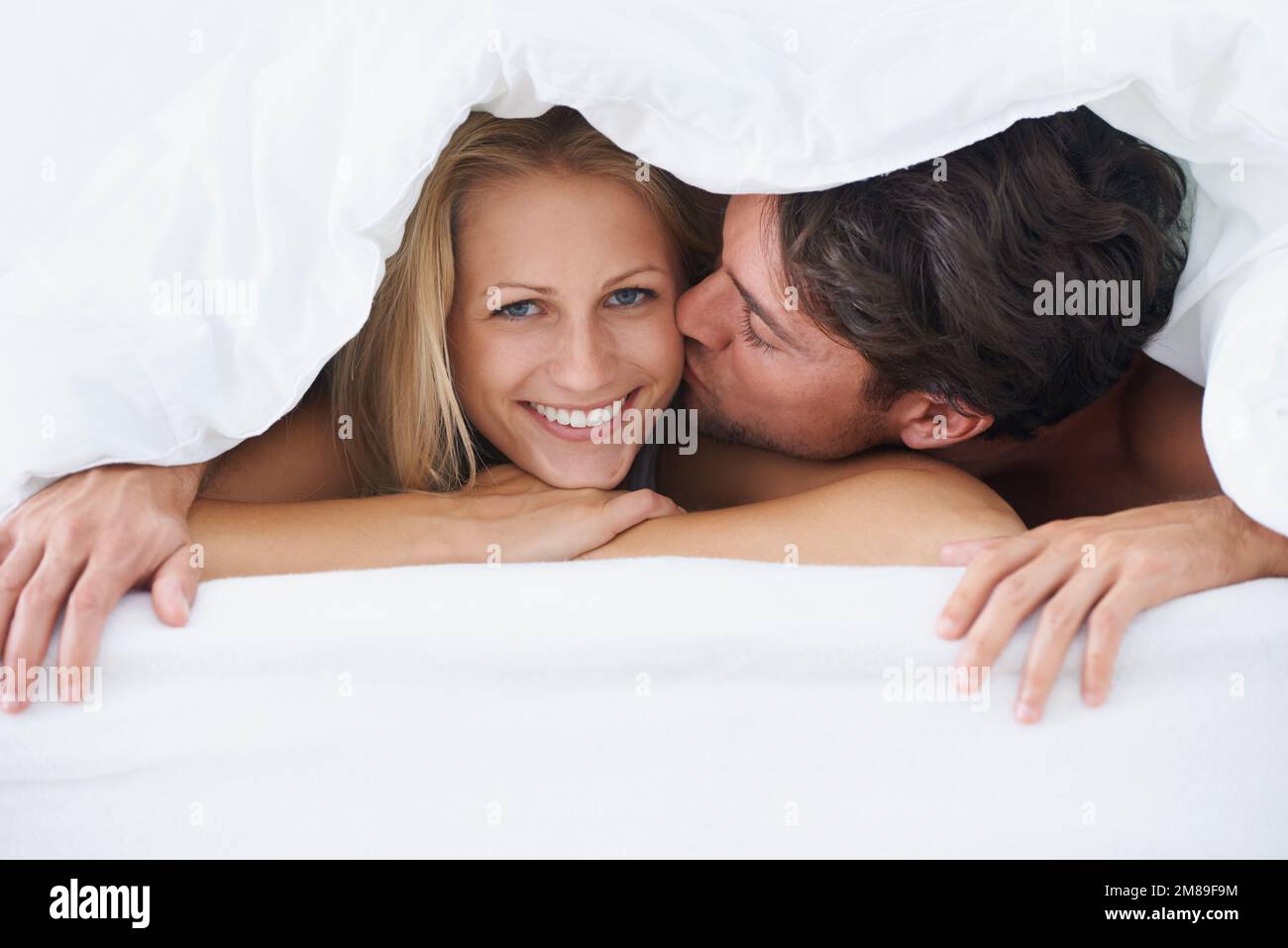 Glücklich mit ihrem Mann. Ein Mann, der eine lächelnde Frau auf die Wange küsst, während er auf einem Bett liegt und unter einem Bettbezug kuschelt. Stockfoto