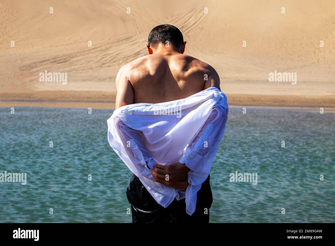 Ein gutaussehender Mann zieht am Meer sein weißes nasses Hemd aus. Muskulärer Rücken Stockfoto