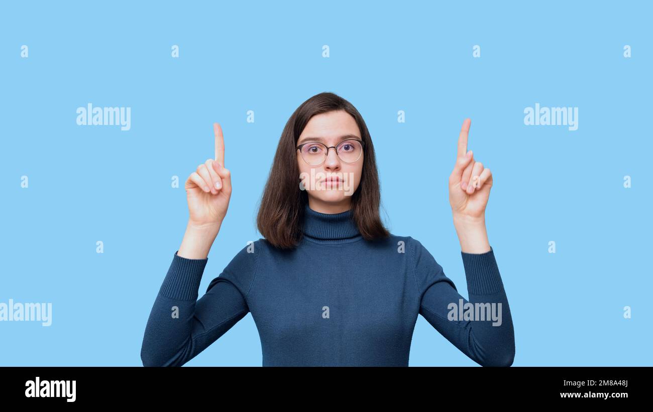 Eine charmante, seriöse Studentin mit Brille in einem blauen Pullover, die mit dem Finger auf einen leeren Raum zeigt, isoliert auf blauem Hintergrund. Das Mädchen zeigt auf Stockfoto
