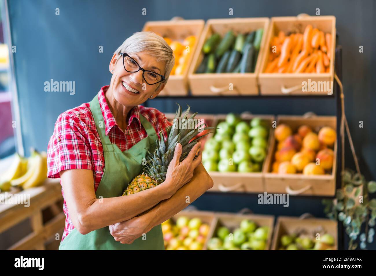 Reife Frau arbeitet im Obst- und Gemüseladen. Sie hat Ananas in der Hand. Stockfoto