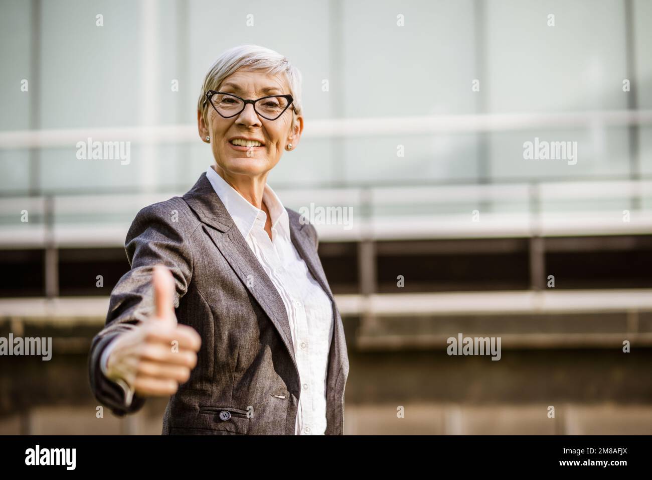 Außenporträt einer erfolgreichen, reifen Geschäftsfrau vor dem Firmengebäude. Sie schaut in die Kamera und zeigt Daumen nach oben. Stockfoto