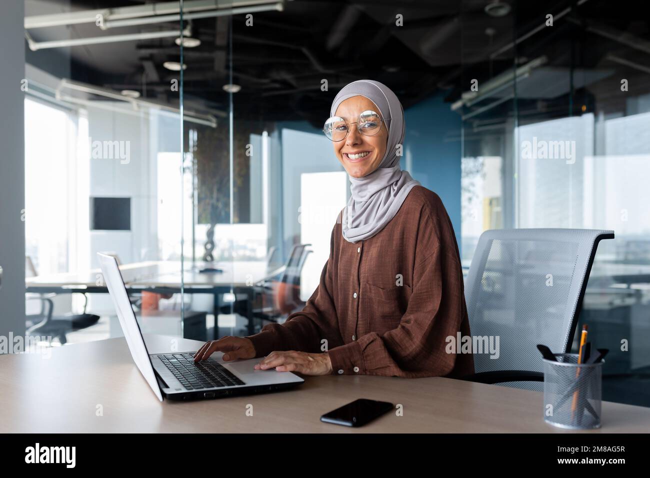 Porträt einer erfolgreichen Geschäftsfrau im Büro mit Laptop, einer Frau im Hidschab, die lächelt und in die Kamera schaut, einer muslimischen Büroangestellten, die eine Brille trägt. Stockfoto