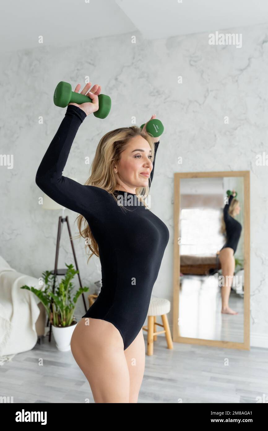 Seitenansicht. Schlanke Frau in schwarzem Body trainiert Hände, Hanteln zu Hause anheben. Blondy, weibliche Fitnesstrainerin, die ein Armtraining macht. Gesunde Körperunterstützung Stockfoto