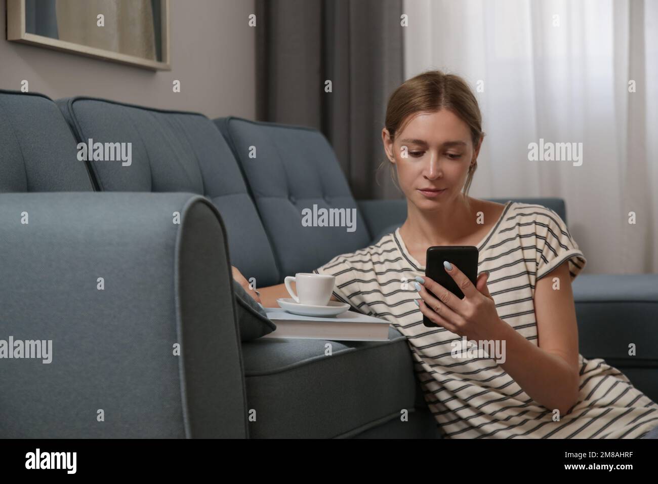 Entspannte junge Frau, die einen entspannten Tag zu Hause mit Telefon, einer Tasse Kaffee und einem Buch genießt Stockfoto