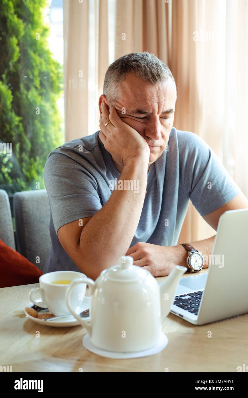 Vertikaler gelangweilter, müder, verwirrter Mann, der im Café an einem Laptop arbeitet, Kaffee trinkt, im Geschäft Geld verdient. Burnout Stockfoto