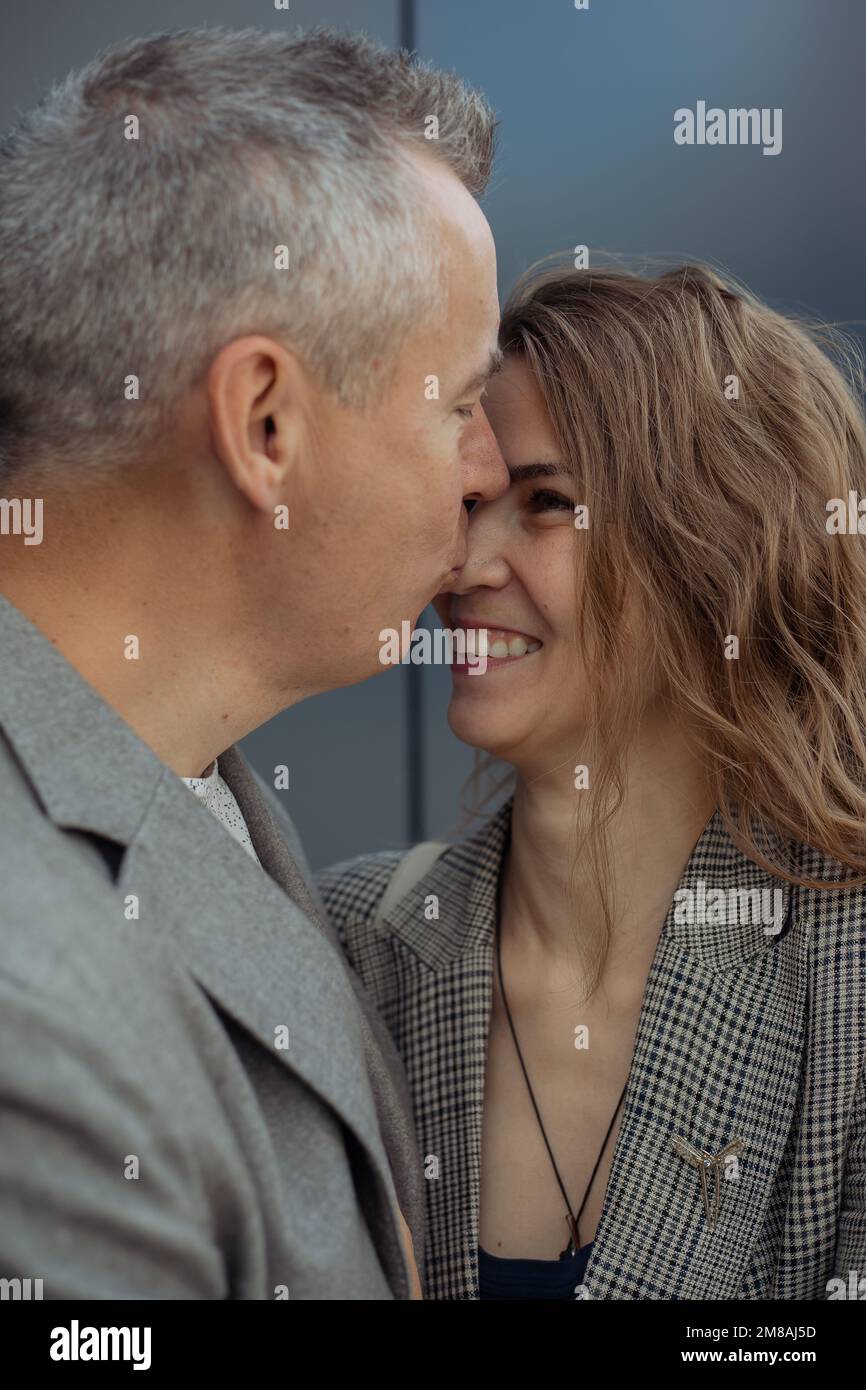 Ein vertikales, romantisches Paar von einem erwachsenen, grauhaarigen Mann, der sich in die Nase küsst, eine hellhaarige, lachende Frau an der grauen Wand. Familie Stockfoto
