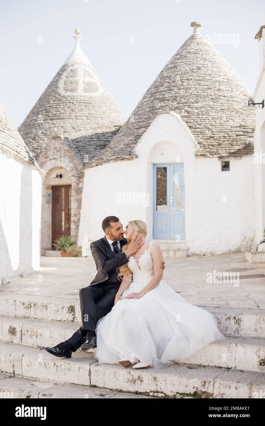 Glückliches Hochzeitspaar, das auf der Treppe in der Nähe von konischen Dächern von Trulli sitzt und sich küsst. Junge Braut umarmt sich mit Bräutigam. Stockfoto