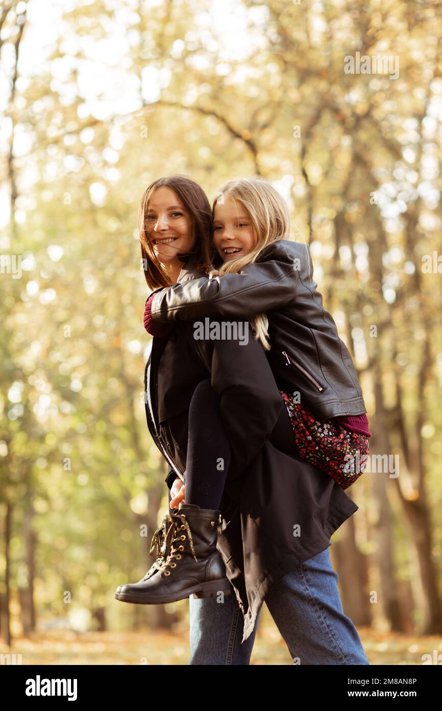 Seitenansicht der Familie von jungen Frauen und Teenagern, die im Wald spazieren. Mutter, die ihre Tochter mit dem Huckepack fährt. Herbst. Stockfoto