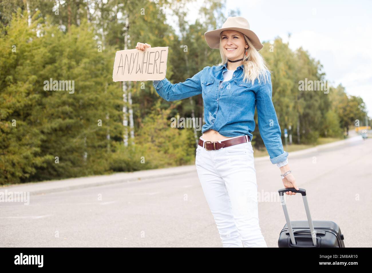 Lächelnd selbstbewusst erlebte glückliche blonde Frau mit Hut trampen mit Karton überall Rollen an Bord Koffer tragen Stockfoto