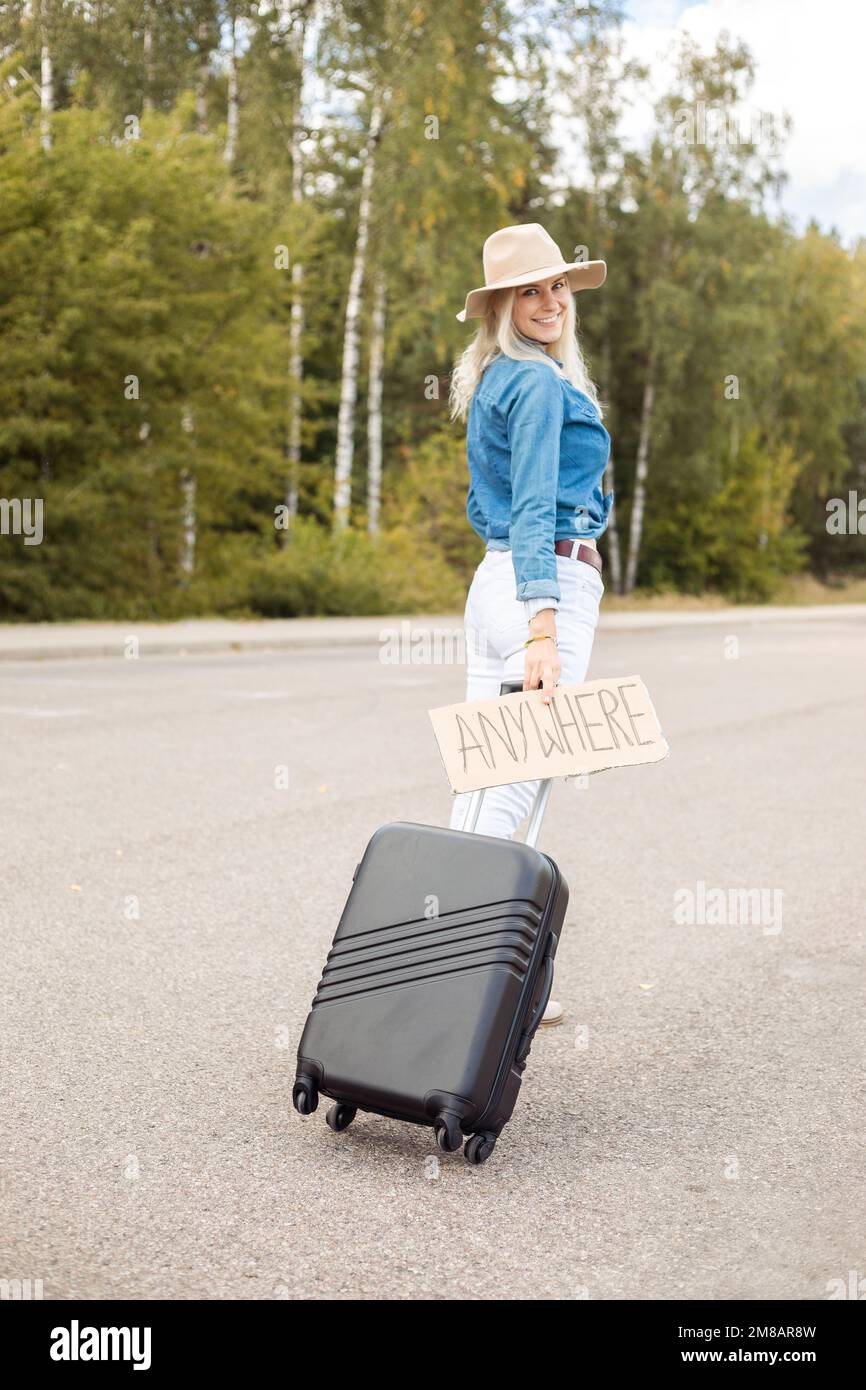 Vertikale lächelnde blonde Anhalter mit Kartonplatten, die Rollen an Bord des Koffers tragen, und wartende Autoanlegestelle Stockfoto