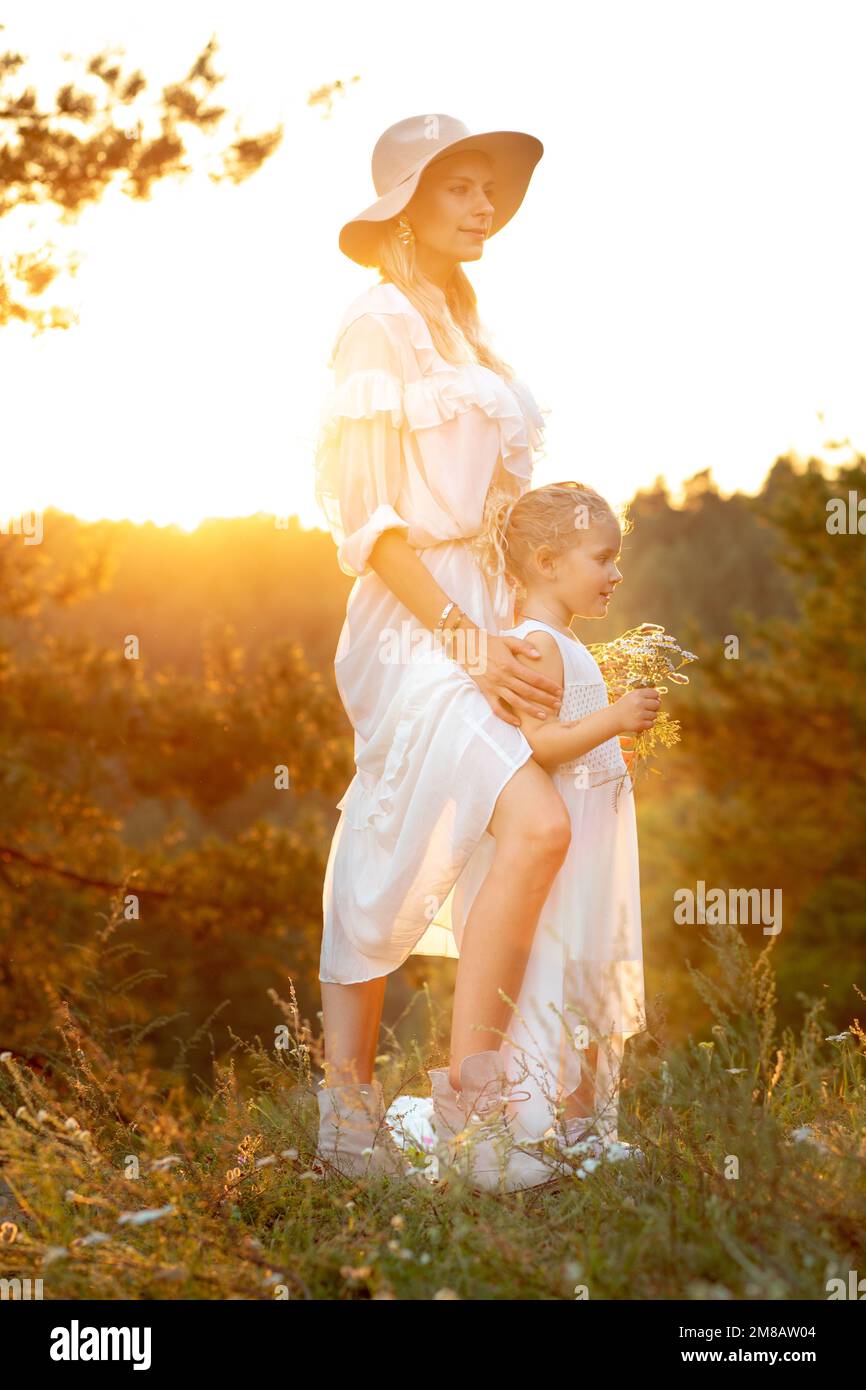 Mutter und kleine Tochter stehen in weißen Kleidern in Nadelholz bei gelbem Sonnenuntergang, die Sonne scheint. Mama und Kind gehen im Sommer abends im Wald spazieren Stockfoto