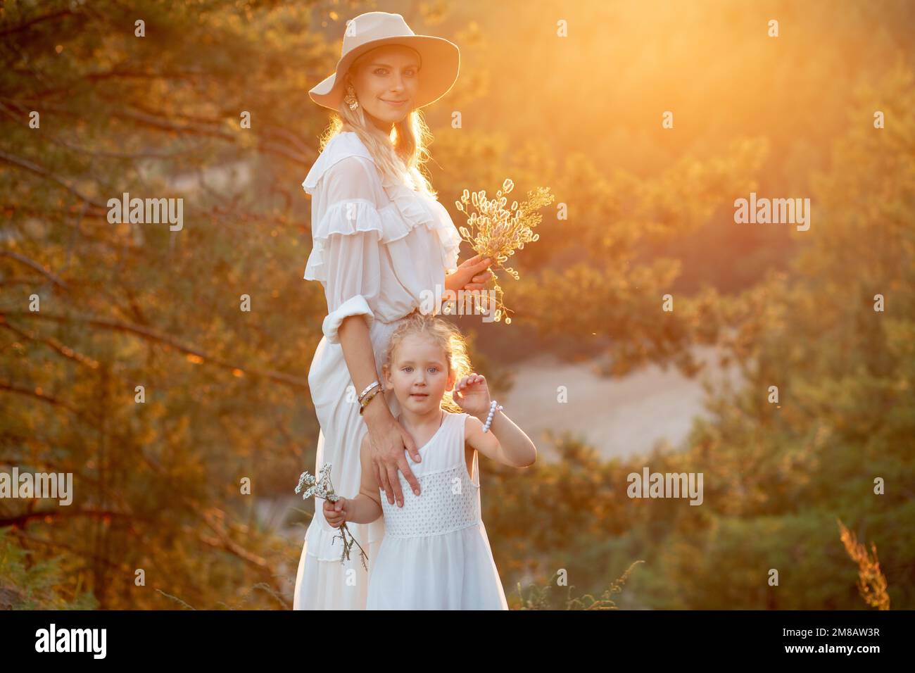 Mutter und süße kleine Tochter stehen auf einem Hügel in weißen Kleidern in Holz bei Sonnenuntergang. Mutter und Kind gehen im Sommer Abend mit im Wald Stockfoto