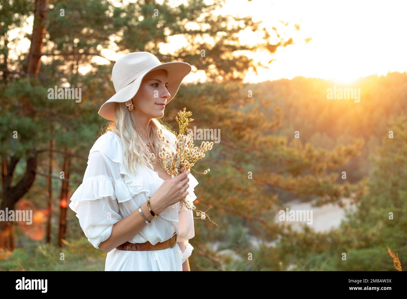 Die schöne Dame steht mit weißer Kleidung und Hut mit einem Blumenstrauß auf einem Hügel in Nadelholz bei Sonnenuntergang. Eine Frau kommt rein Stockfoto