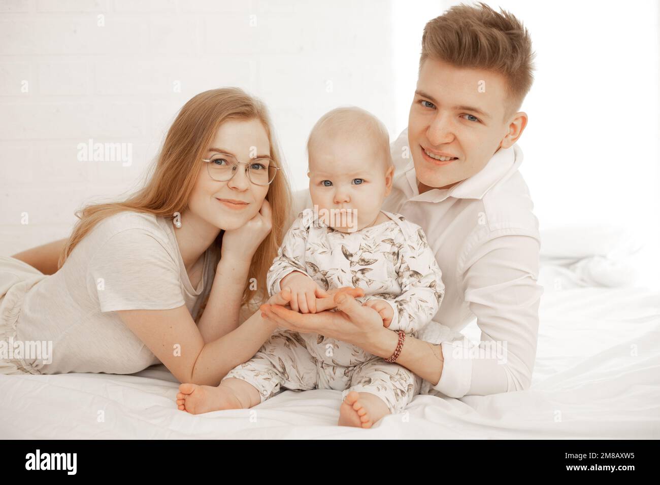 Ein glückliches junges Paar mit einem kleinen Baby liegt auf dem Bett und hält sich die Handflächen aneinander, weißer Hintergrund, kostenloser Kopierbereich. Familienfoto von mom, Dad und Stockfoto