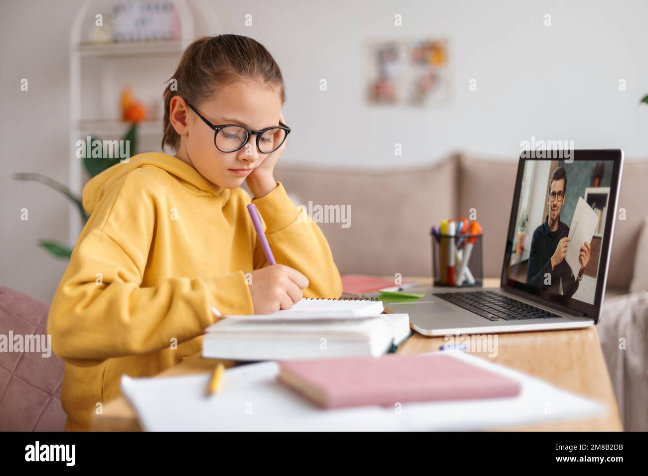 Ein müdes Mädchen mit Brille, das Online-Unterricht mit einem Privatlehrer auf dem Laptop hat. Der Junge langweilte sich vom Lernen, schrieb Mathe-Übungen in Notizbücher. Seitenansicht. E-Learning und Fernschulen während der Covid-Quarantäne Stockfoto
