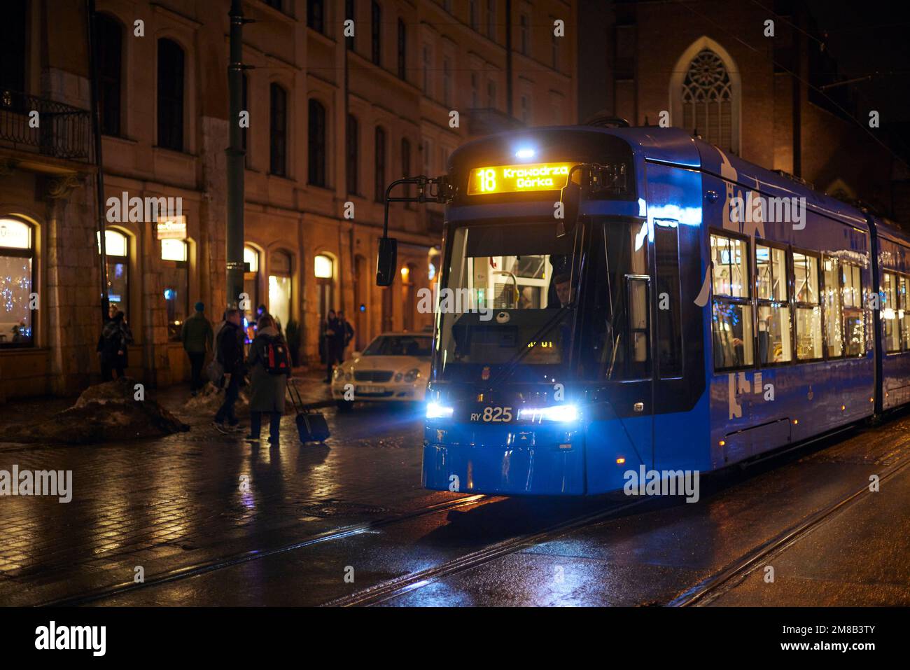 Blaue Straßenbahn am Abend in Krakau mit beleuchteten Fenstern und Menschen. Polnische Architektur, orangefarbenes Licht aus Laternen und Reflexionen. Stockfoto