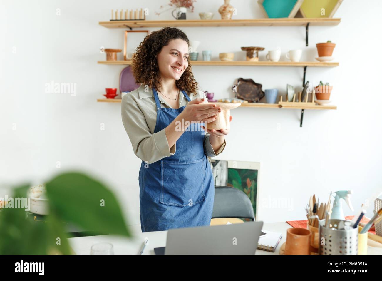 Junge Geschäftsfrau in ihrer Werkstatt lächelt vor dem Hintergrund von keramischen Produkten. Konzept Start-up für kleine Unternehmen. Stockfoto