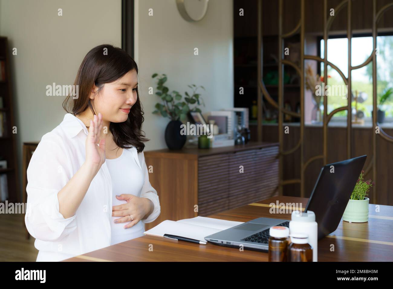 Asiatische Schwangere, die eine Pillenflasche hält, kommuniziert mit dem Arzt im Internet im Wohnzimmer zu Hause. Telemedizin, Schwangerschaft, Baby erwarten und gesund Stockfoto