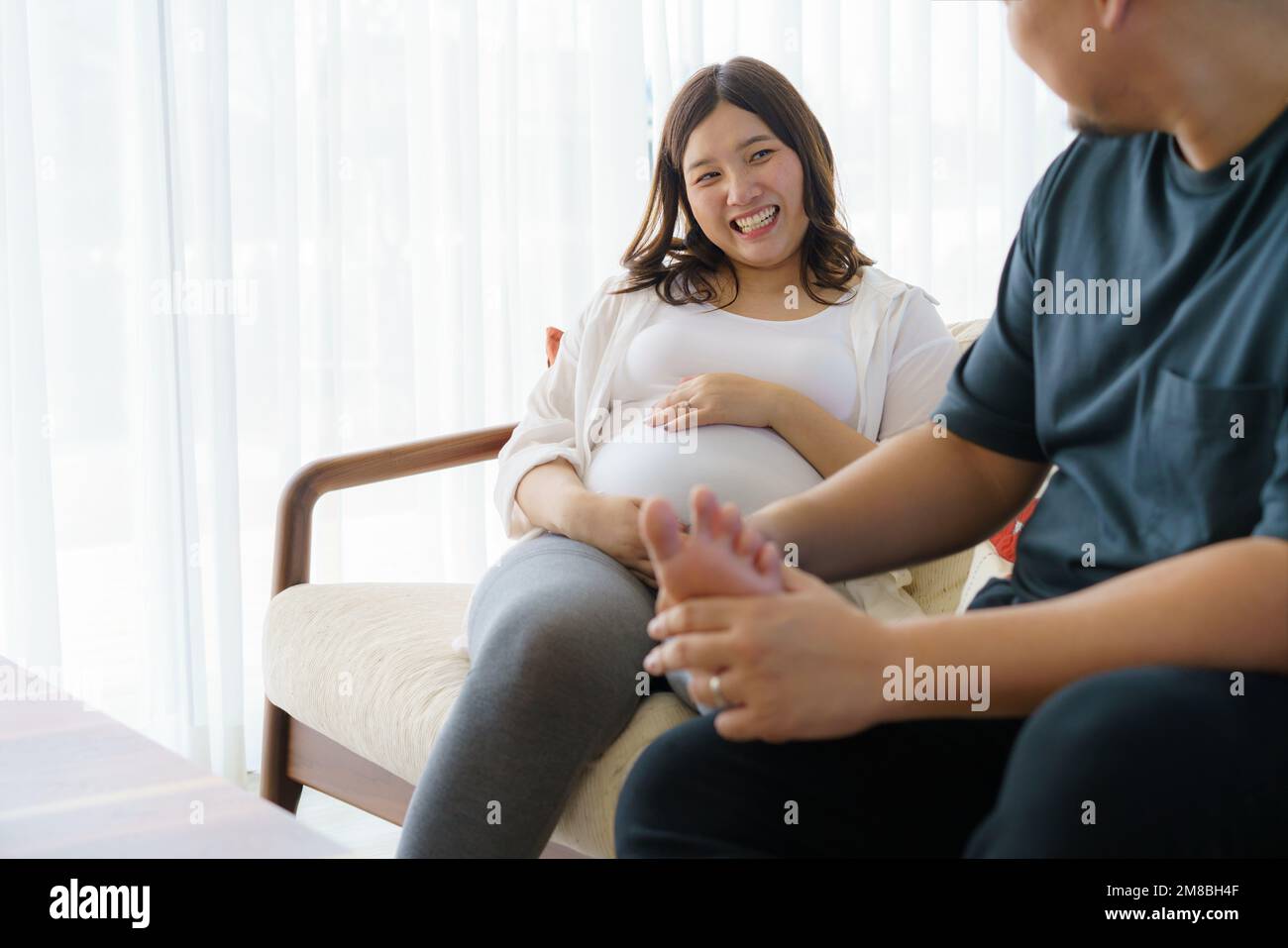Der asiatische Ehemann massiert die Füße der schwangeren Frau im Wohnzimmer des Hauses. Glückliche schwangere Familie. Stockfoto