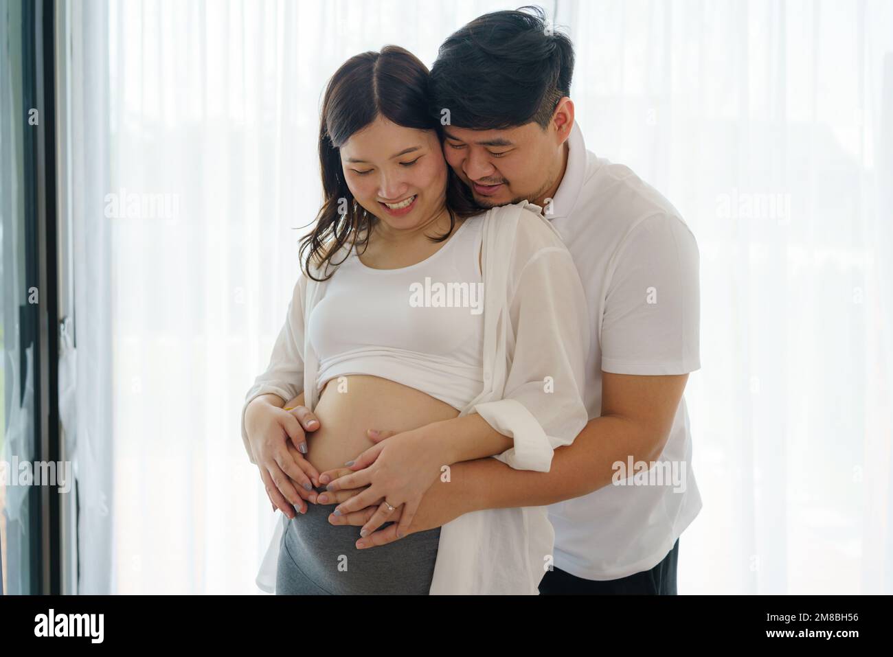 Glückliches asiatisches Paar erwartet ein Baby, das zu Hause am Fenster steht, liebevoller Ehemann, der zärtlich den Bauch seiner schwangeren Frau berührt Stockfoto