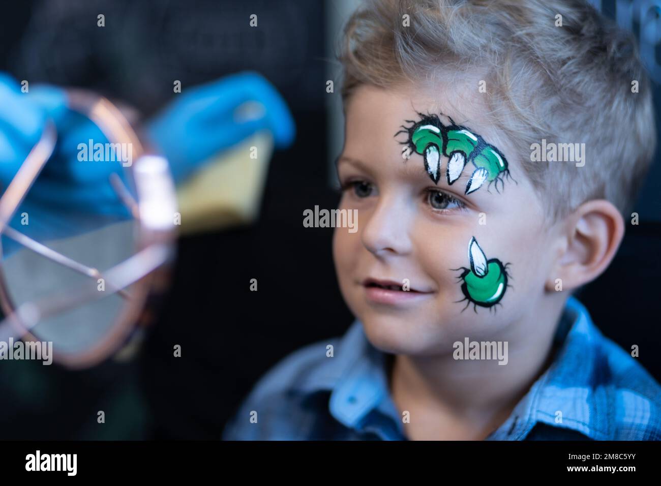 Auf einer Kinderparty sieht ein süßes Kind ein Bild auf seinem Gesicht im Spiegel – ein Bild von Dinosaurier- oder Drachenkrallen. Stockfoto