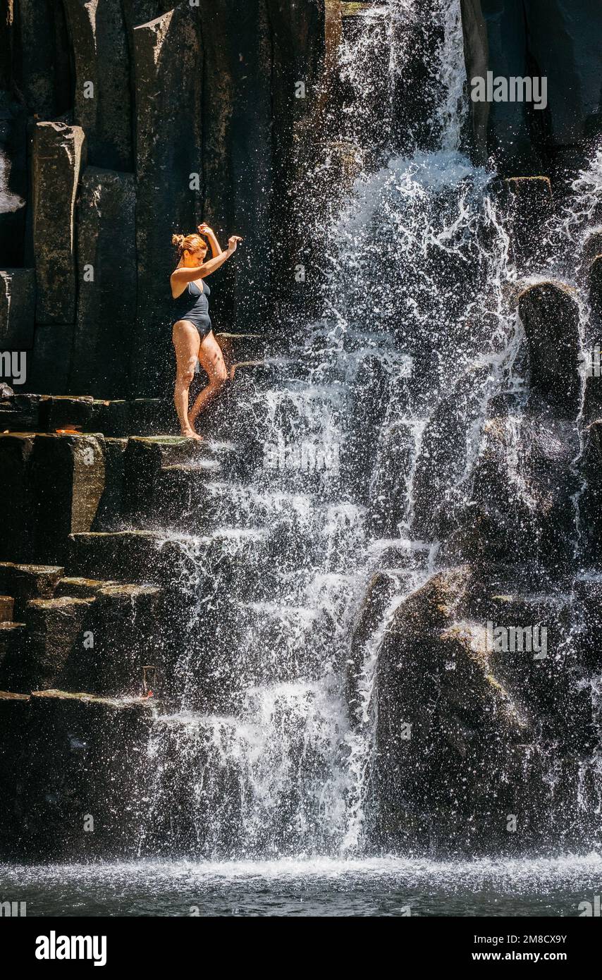 Weiße Frau in schwarzem Badeanzug erfrischend unter den Wasserströmen, die über schwarze Vulkansteinkaskaden fließen. Wasserfall Rochester Falls – Pop Stockfoto