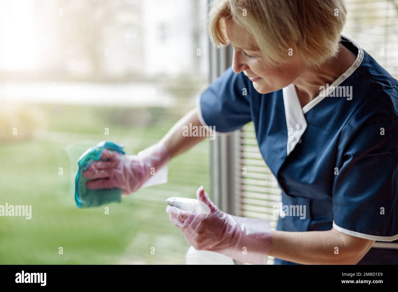 Leitende Zimmermädchen in Handschuhen und einheitliches Reinigungsfenster mit Lappen und Reinigungsspray Stockfoto