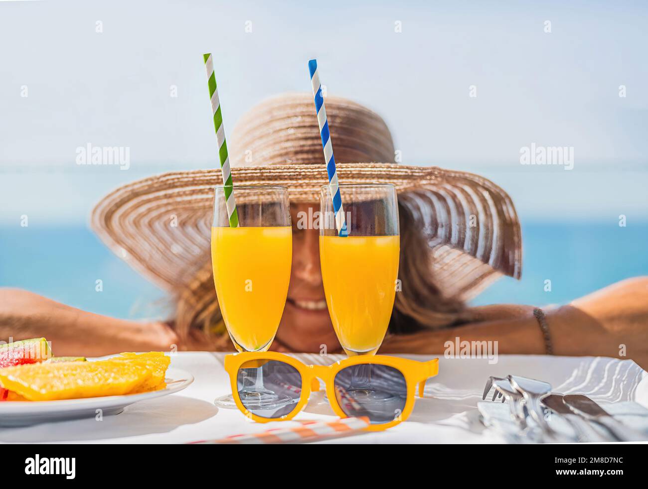 Nahaufnahme von Orangensaftgläsern und gelber Sonnenbrille, vor einer Frau mit Strohhut im Hintergrund unscharf. Blaues Meer im Hintergrund. Urlaub Stockfoto