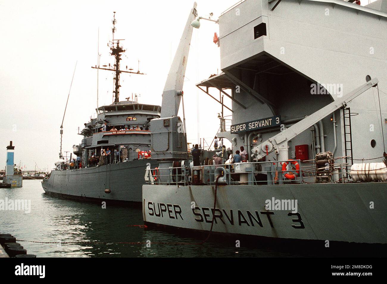 Der Meeresminenräumer USS LEADER (MSO-490) ist über dem Unterwasserdeck des niederländischen Schwertransportschiff SUPER SERVANT 3 gesichert. Drei US-Dollar Minenräumer der Marine und ein Minenabwehrschiff werden an Bord des SUPERDIENERS 3 geladen, um zum Persischen Golf zu transportieren, wo sie US-Streitkräfte unterstützen werden, die als Reaktion auf die Invasion Kuwaits durch den Irak in die Region geschickt werden. Der nuklearbetriebene Flugzeugträger USS ENTERPRISE (CVN-65) befindet sich im Hintergrund. Betreff Betrieb/Serie: WÜSTENSCHILD/STURMSTÜTZPUNKT: Marineflugstation, Norfolk Bundesstaat: Virginia (VA) Land: Vereinigte Staaten von Amerika (USA) Stockfoto